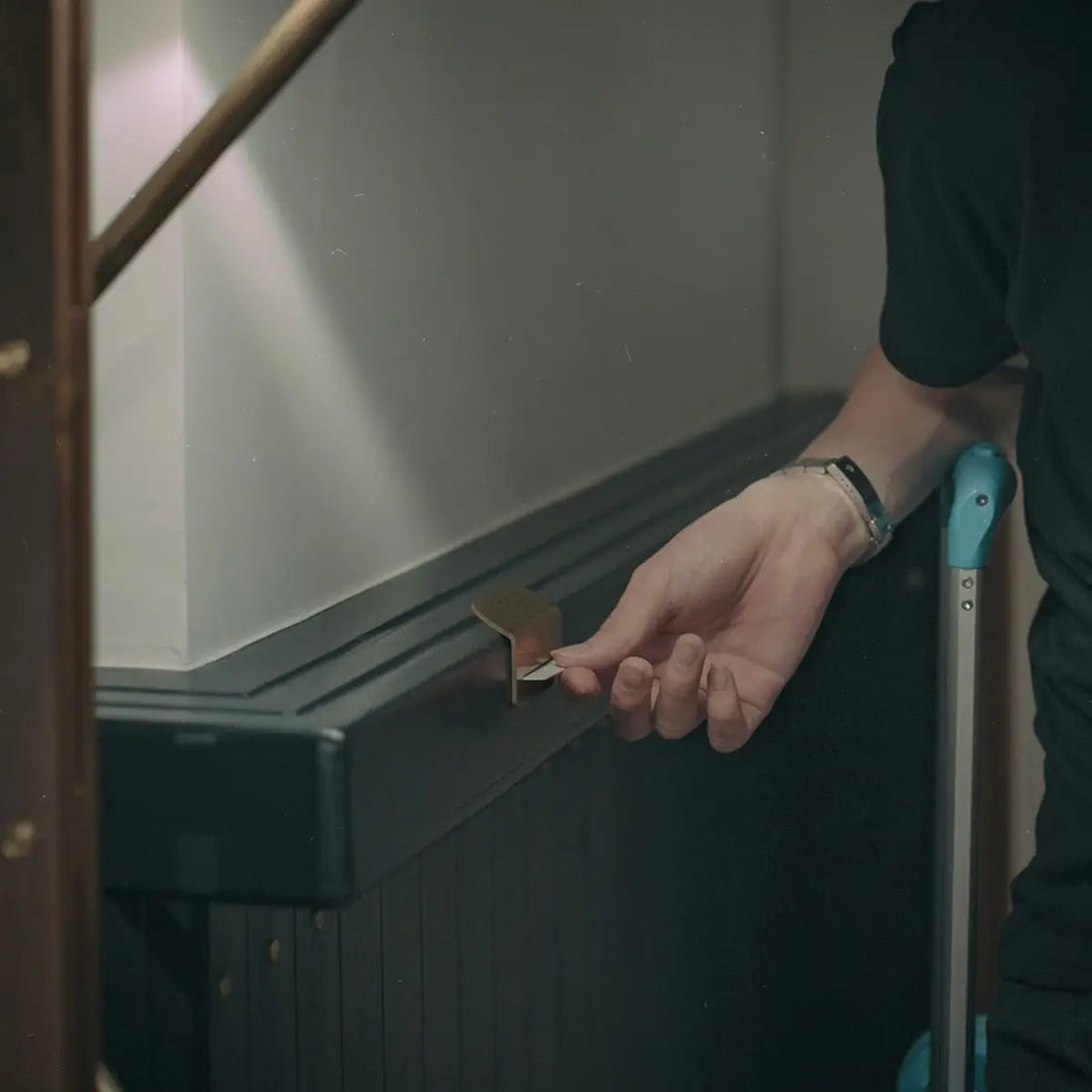 En persons hand håller i ett litet, metalliskt, vinklat nyckelliknande föremål, möjligen för att öppna eller justera något på en svart, lamellförsedd barriär eller möbel.